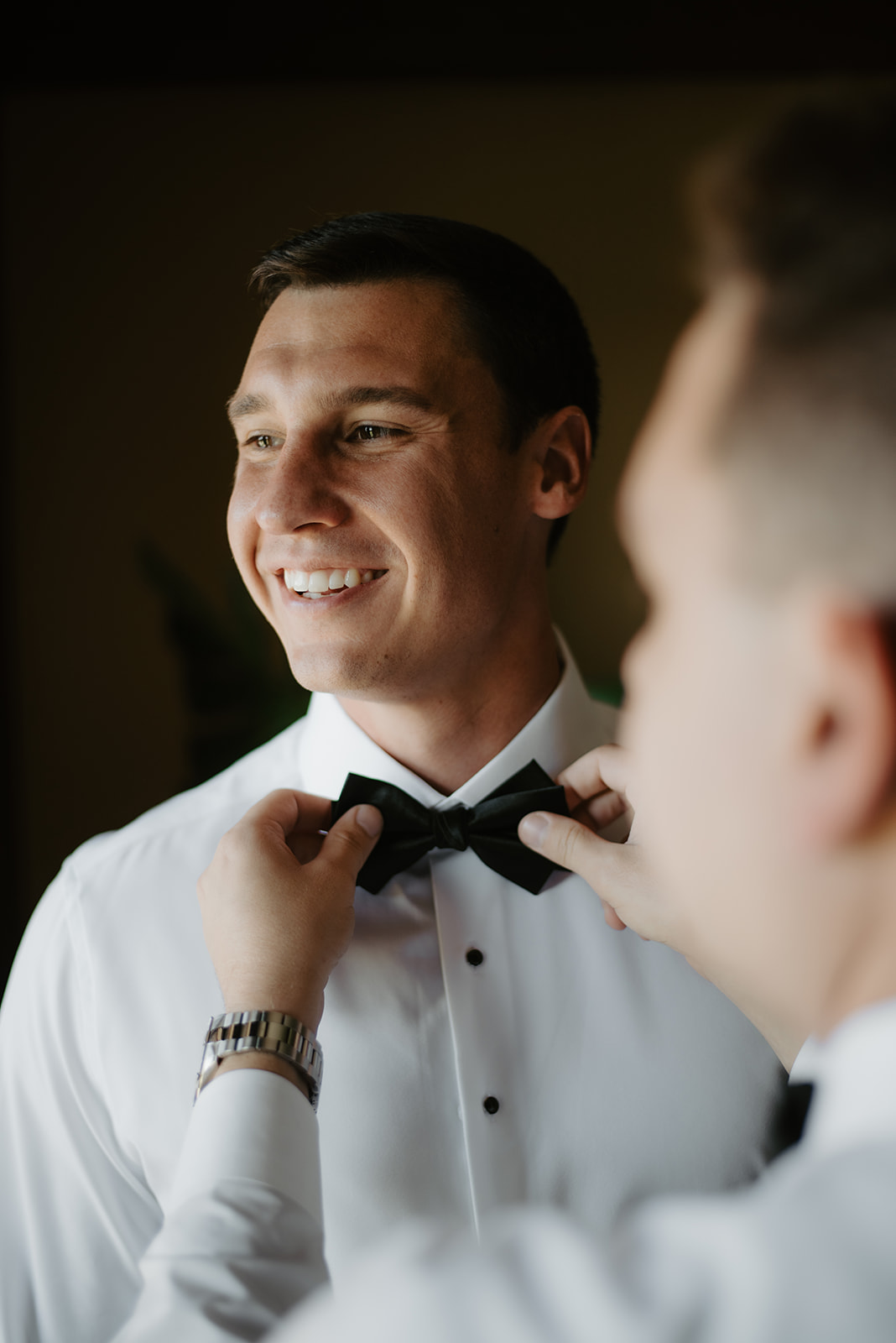 Groom's friend adjusting the groom's black bow tie as he smiles during wedding prep.